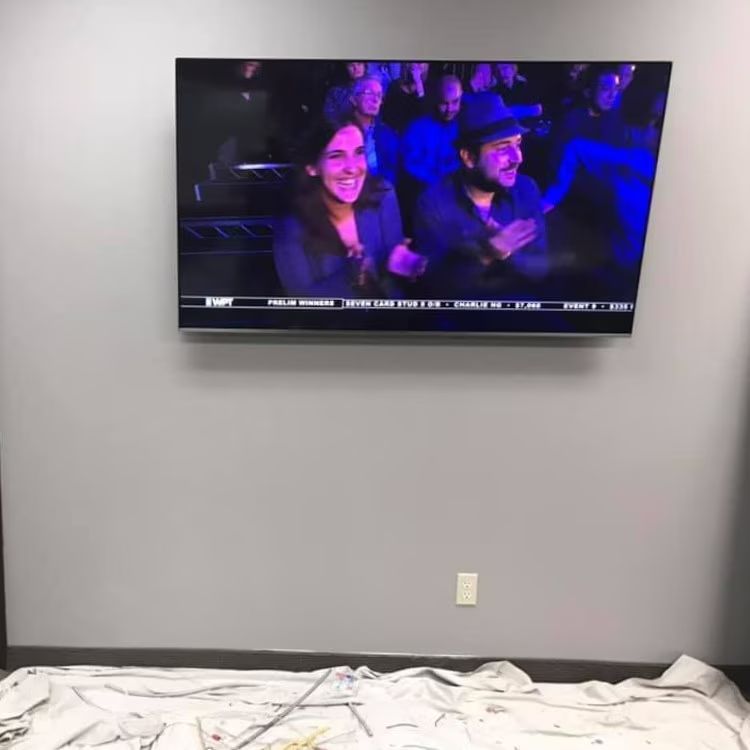 Wall-mounted TV showing a live audience; bed edge in foreground against a gray wall.