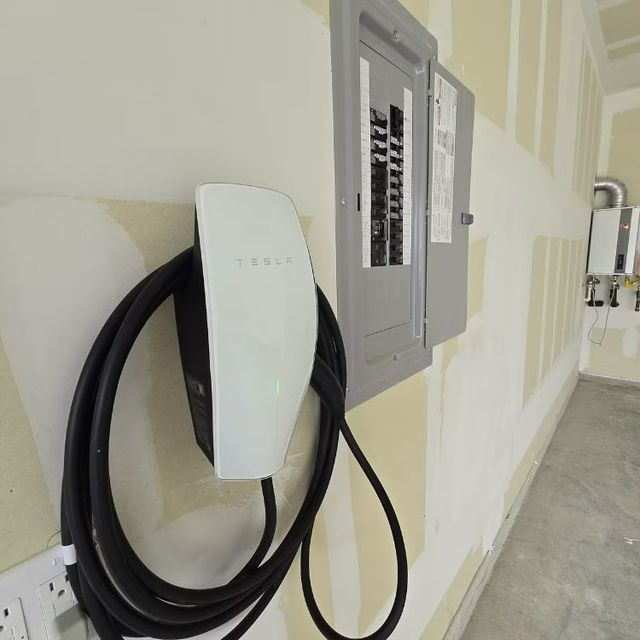Wall-mounted EV charger with coiled black cable beside an electrical panel in a hallway