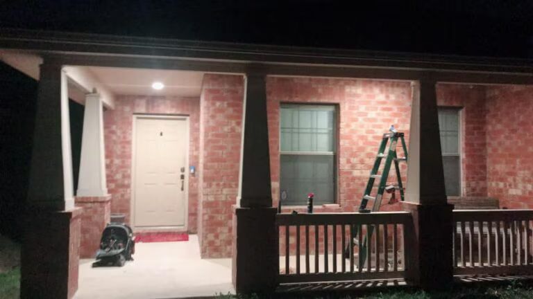 Front porch of a brick house at night, with a ladder on the railing and a closed front door.