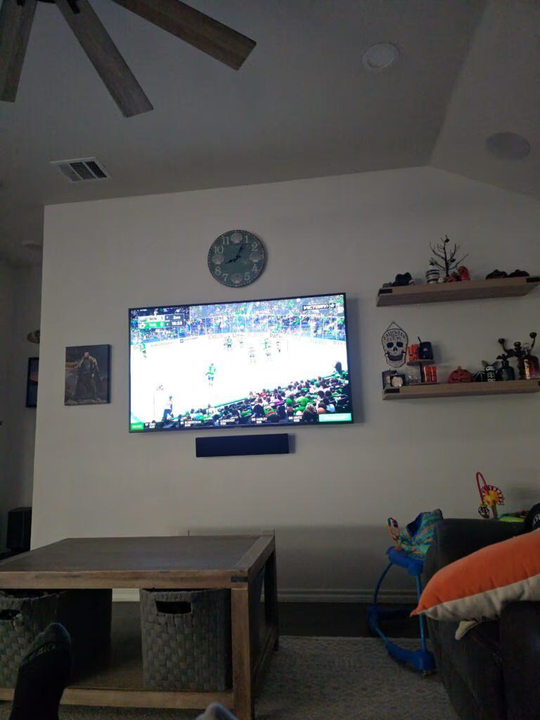 Living room with wall-mounted TV showing a sports game, clock above, shelf decor, and a coffee table.