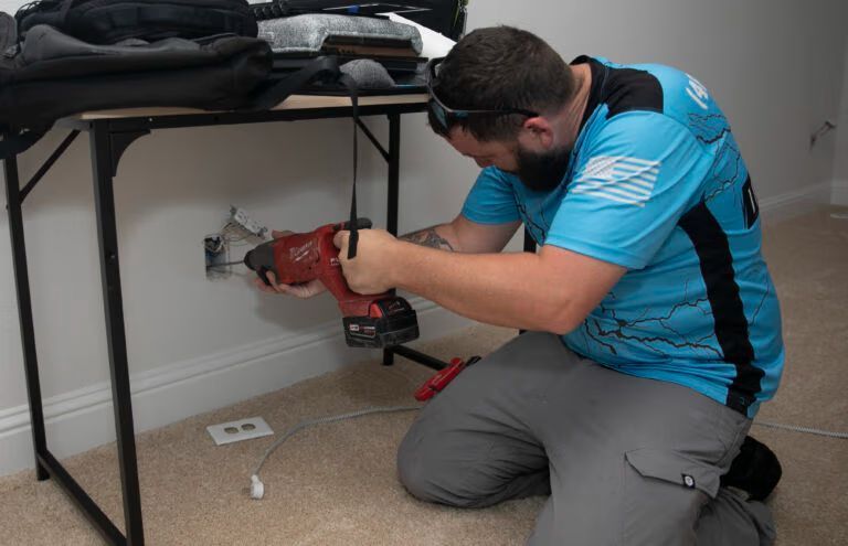 Man kneels under a table, using a red power tool to drill into the wall near an outlet.
