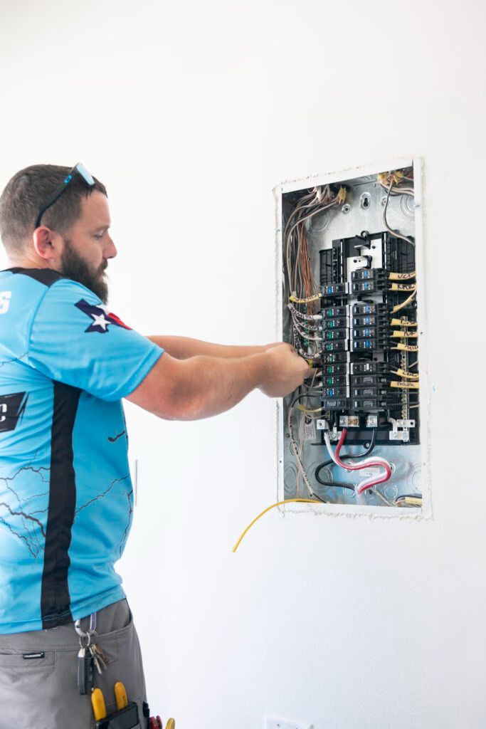 Electrician in blue shirt works inside an open wall electrical panel.