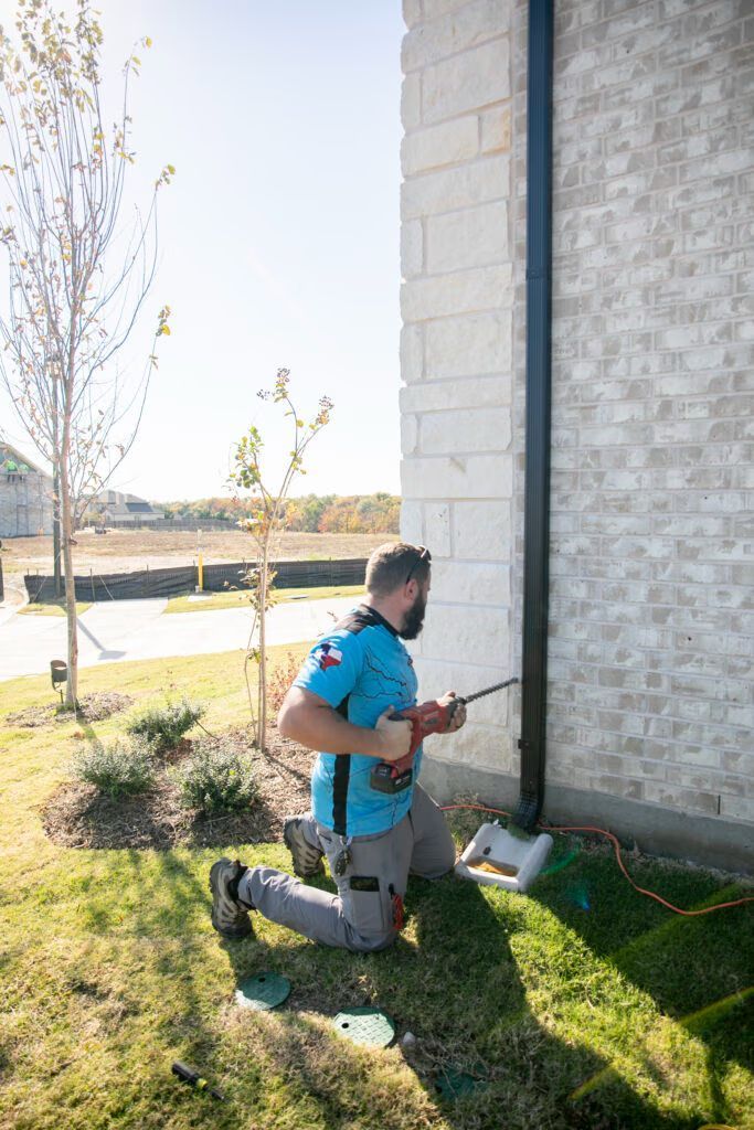 Person kneels beside a white brick wall outdoors, holding a tool near a small utility box in a grassy yard.
