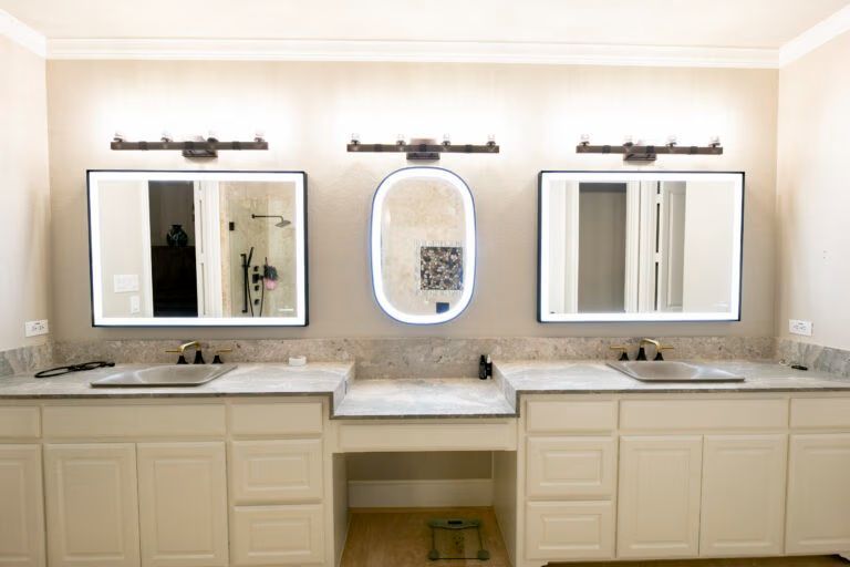Bathroom vanity with three mirrors, double sinks, and beige cabinetry under warm wall lights