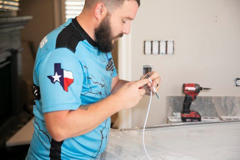 Man in blue work shirt inspecting an electrical component in a room with a drill on the counter