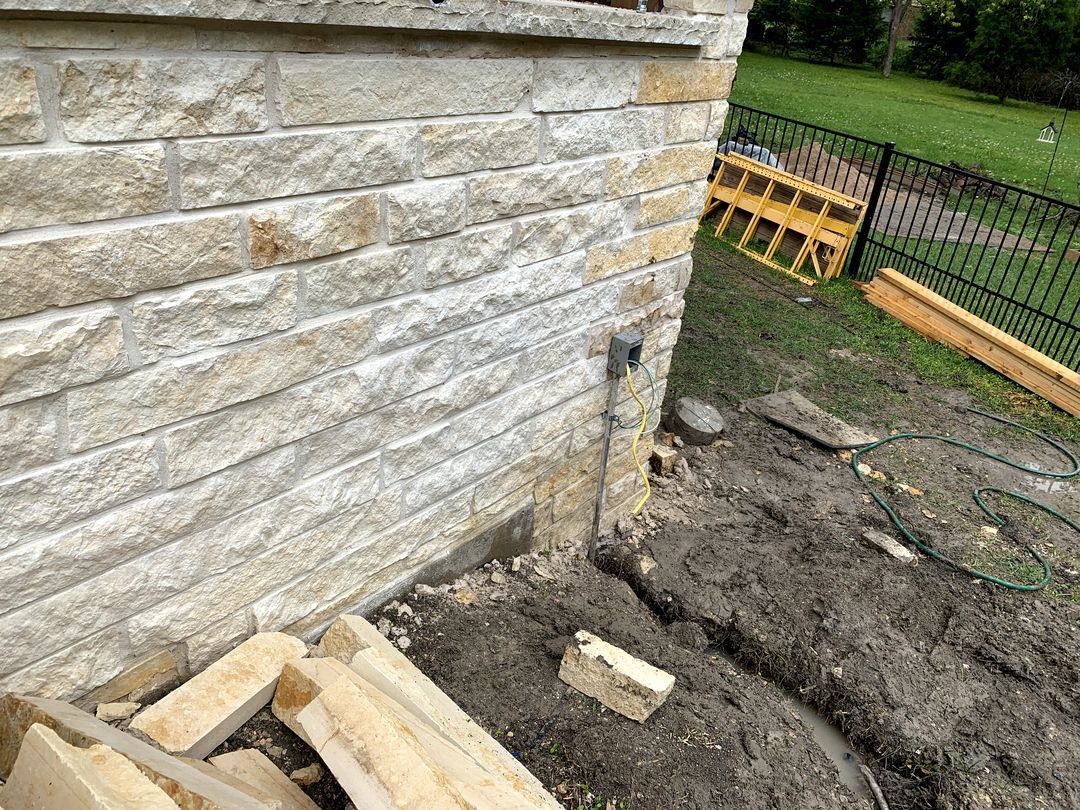 Stone house wall beside muddy ground with a trench, wood scraps, and a fence in the background