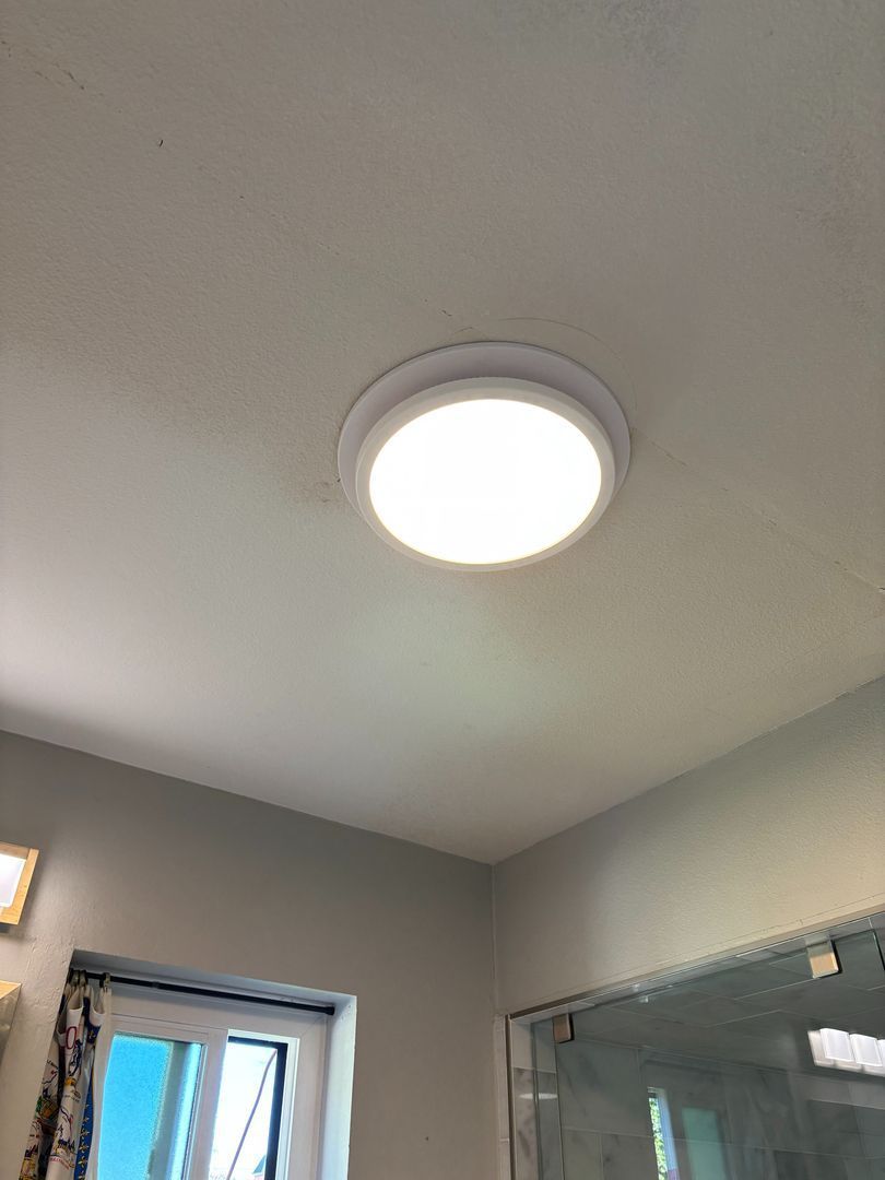 Bright round ceiling light in a gray bathroom with a window and glass shower enclosure.