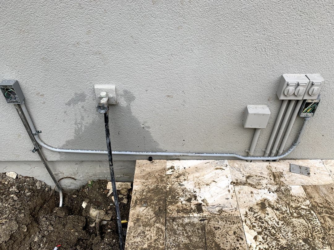 Exterior wall with electrical conduit and utility boxes beside a dirt trench and concrete slab