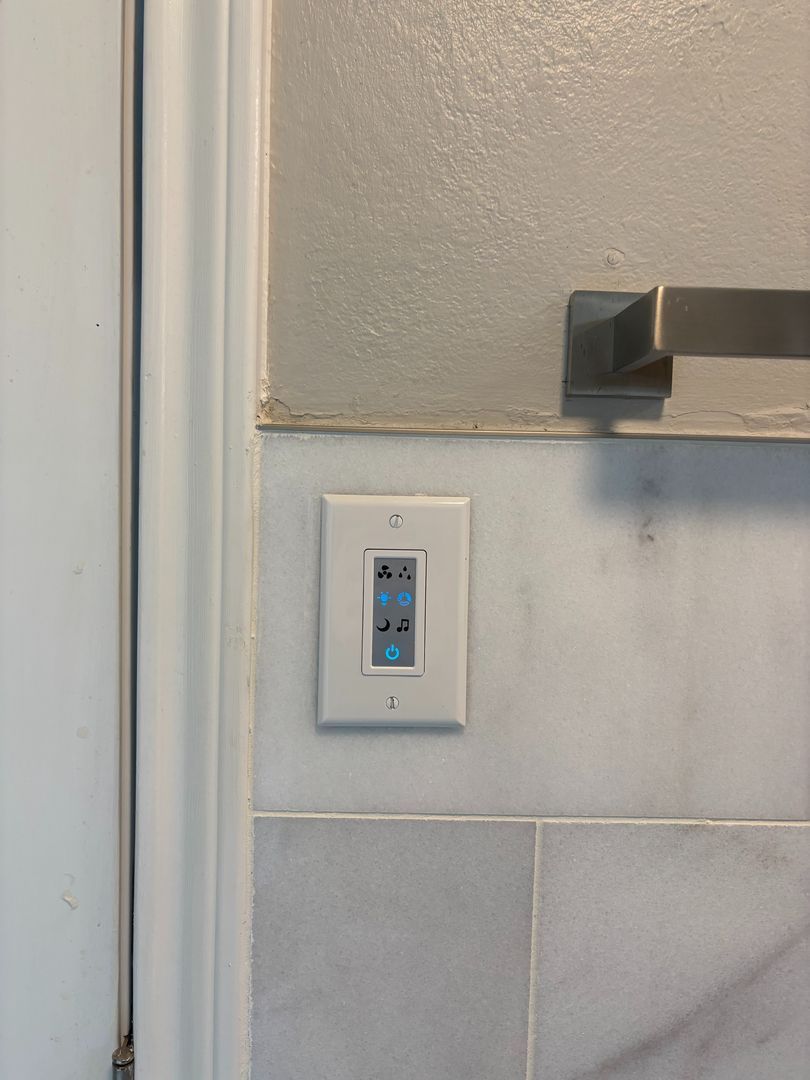 Wall-mounted light switch with blue indicator lights beside a door frame and towel bar.