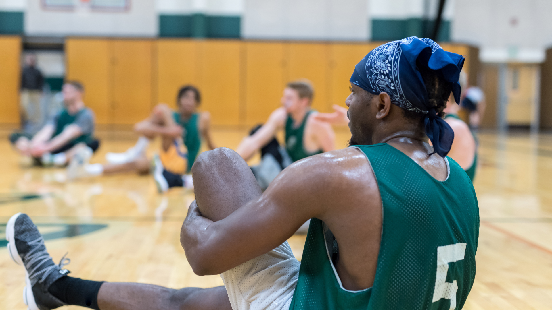 A man in a green jersey with the number 5 on it is stretching on a basketball court.