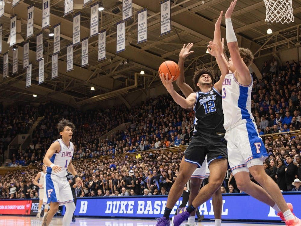 Basketball player in blue jersey shoots over opponent in arena, crowd in background.