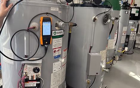 Row of gray industrial water heaters with gauges, labels, and orange temperature meters in a utility room