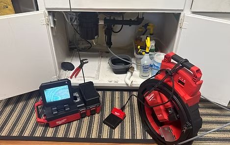 Plumbing drain inspection setup with red sewer camera equipment under a sink cabinet.