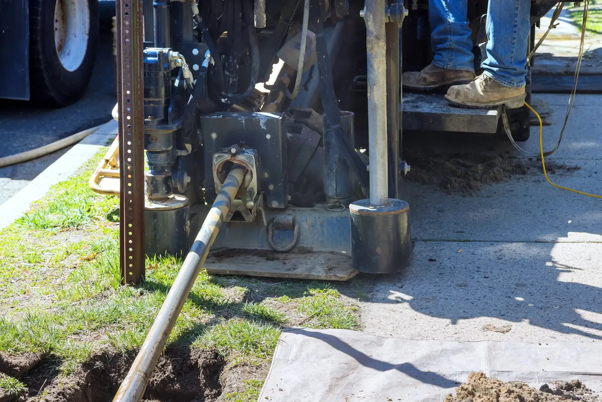 A long metal pipe connected to a machine on a construction site, with workers’ legs visible nearby.