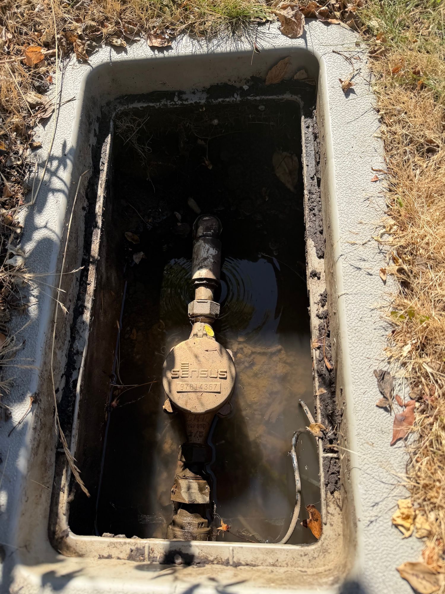 Open utility box with a water shutoff valve and pipe inside, surrounded by dry grass and dirt.
