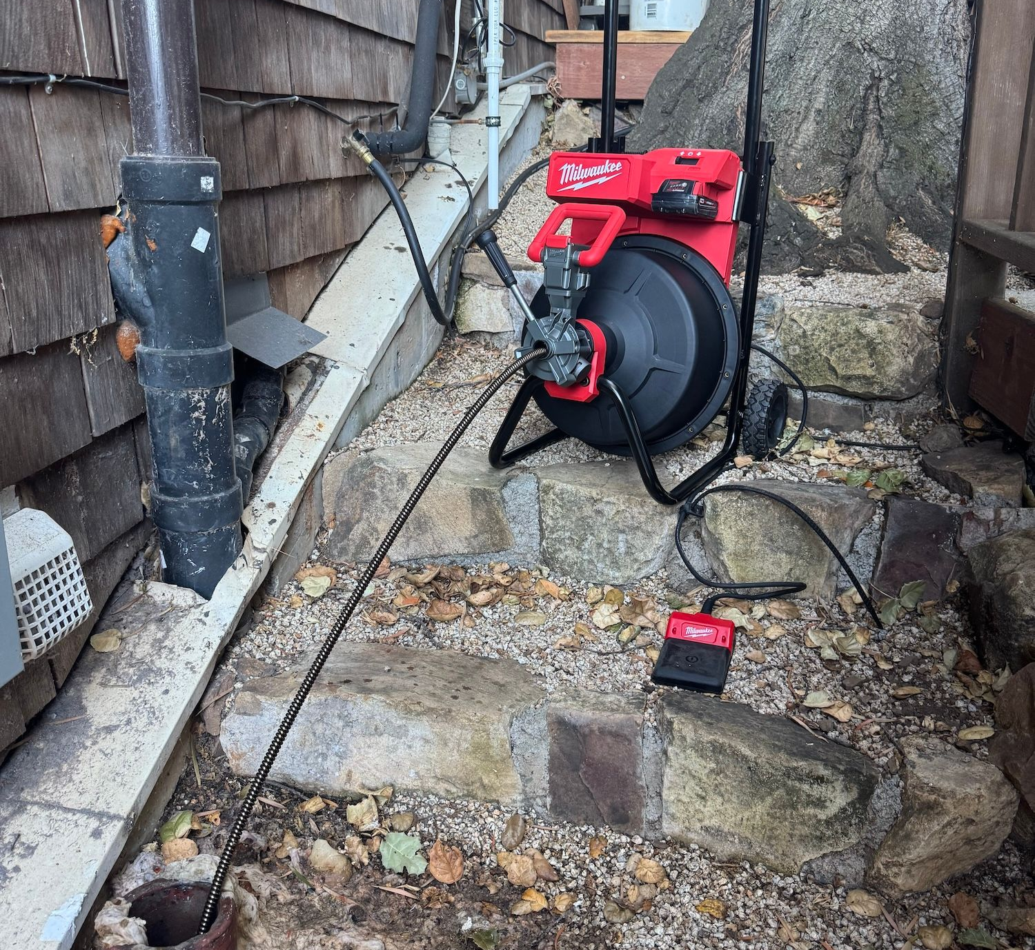 Red hose reel on a rocky path beside a house, with a hose leading to a drain pipe.