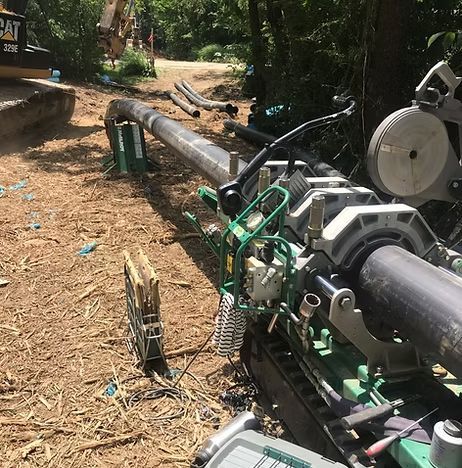 A large black pipeline being laid through a wooded dirt path by green machinery.