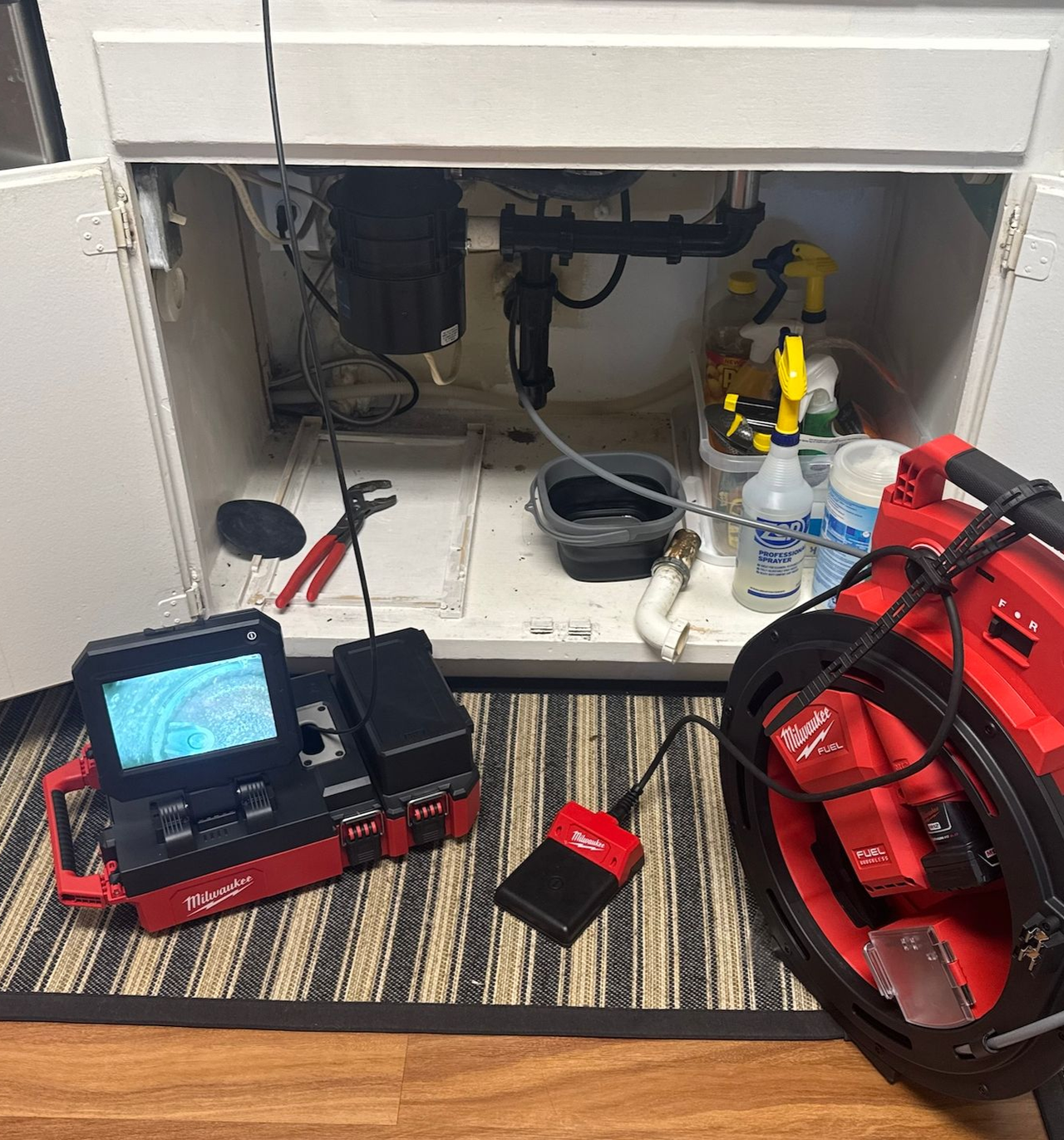 Open cabinet under sink with plumbing tools, red drain camera, and coiled cable on a striped rug