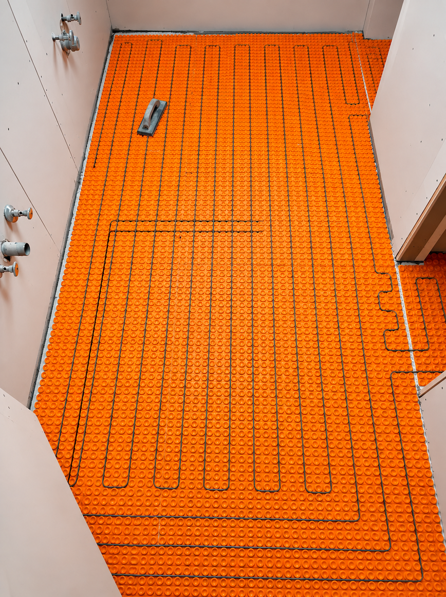 Orange underfloor heating pipes installed on a floor between white walls, with a small tool nearby