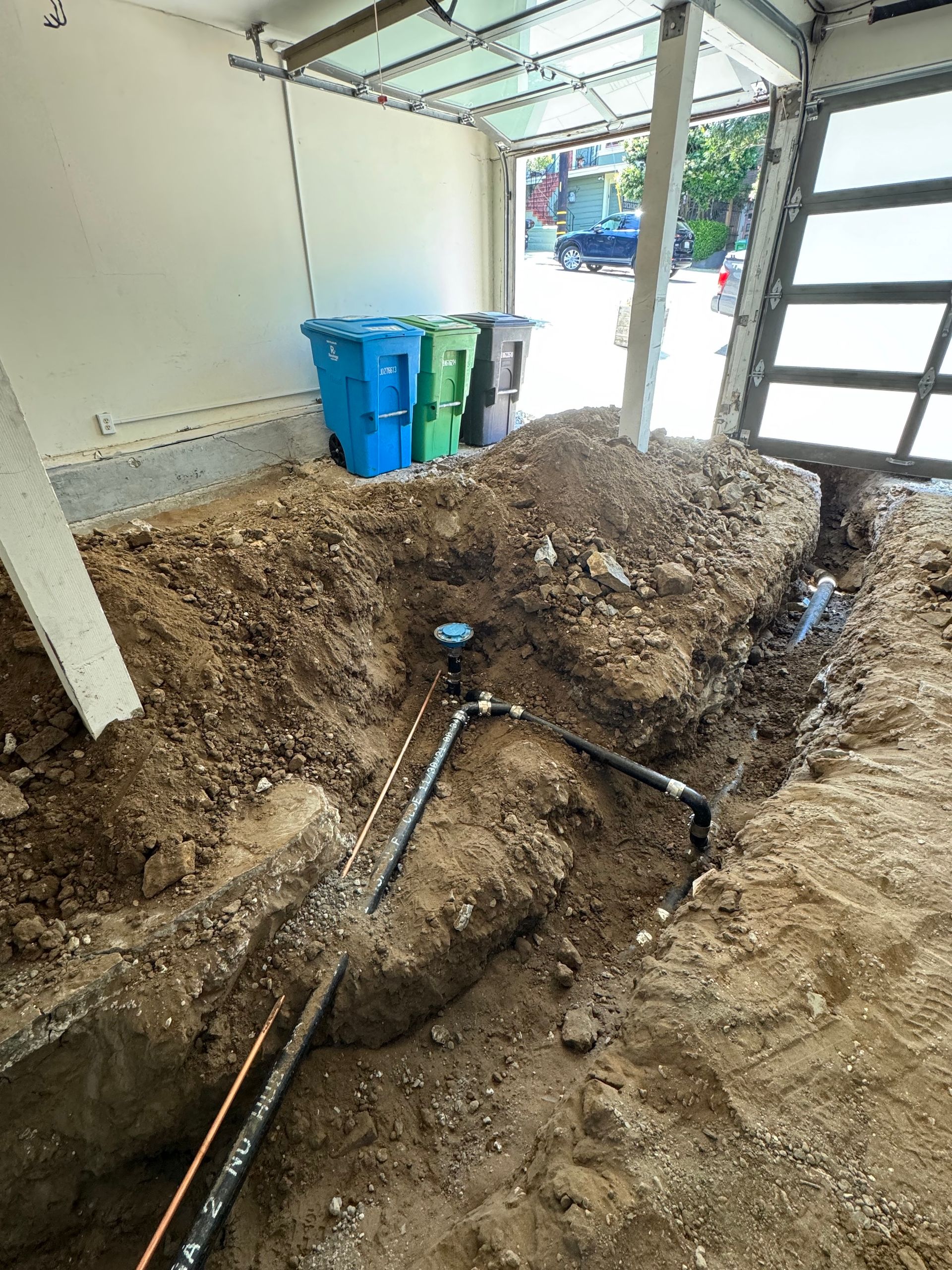 Piles of dirt over exposed pipes in a garage, with trash bins and an open door in the background
