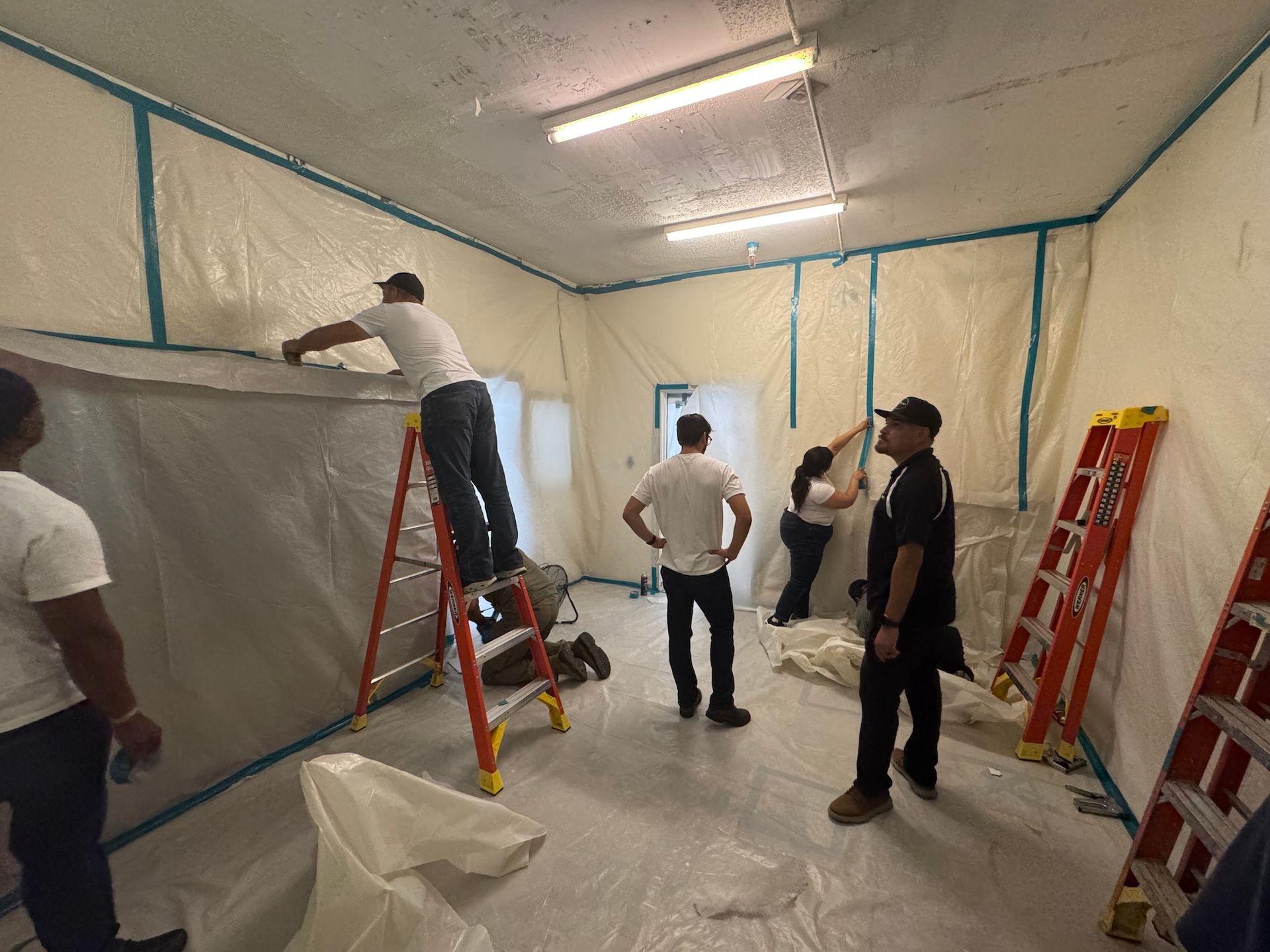 Workers painting and taping a room under renovation, with ladders and protective sheeting.