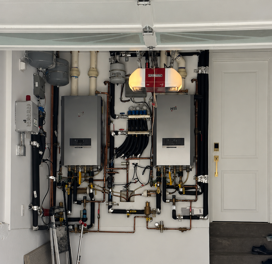 Wall-mounted boiler and water heater system in a utility room with pipes, gauges, and electrical panels