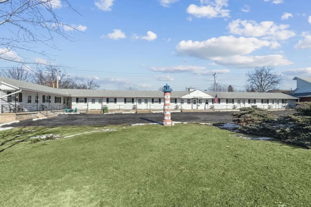 Single-story building with a grassy courtyard and a brick post under a partly cloudy sky.