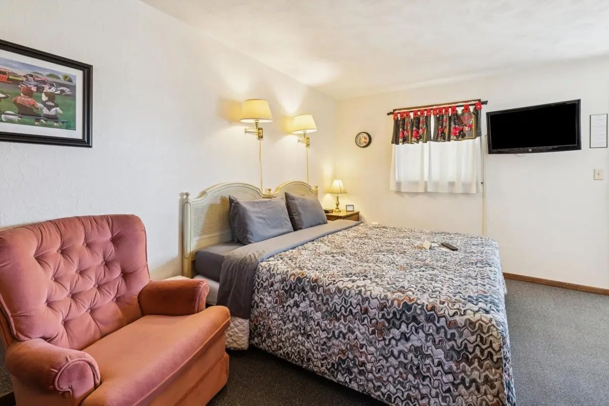 Cozy bedroom with pink armchair, bed, lamps, wall art, and a small TV on the wall.