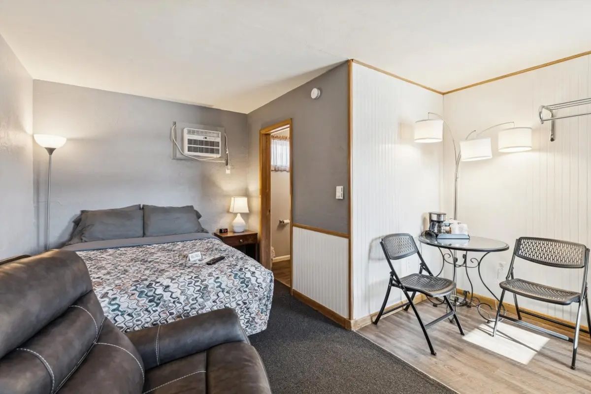Small hotel room with bed, armchair, side tables, and a sitting nook with two chairs and a lamp.