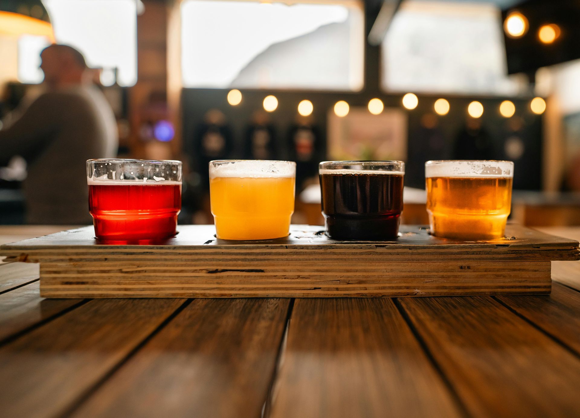 Four small beer glasses on a wooden board at a bar, with blurred lights in the background