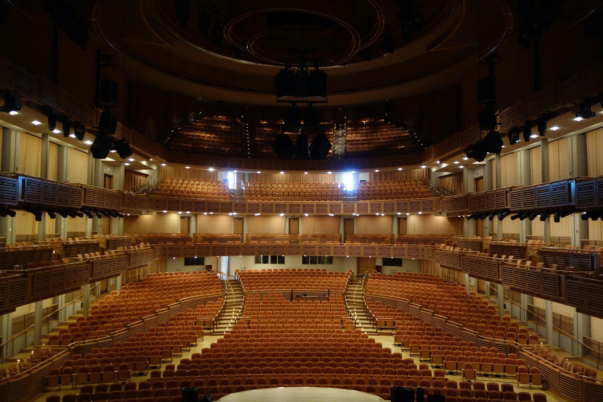 Empty ornate theater auditorium with rows of orange seats and a balcony under warm lights