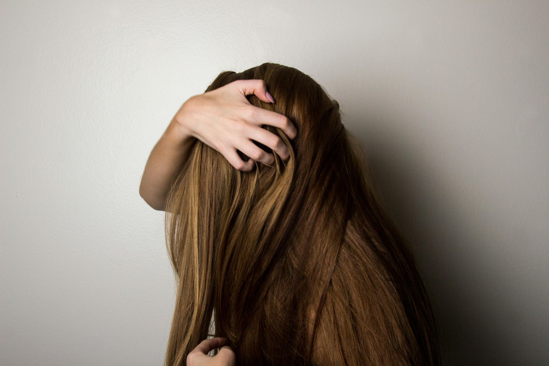 Woman with long, brown hair, hand in hair, against a plain gray wall.