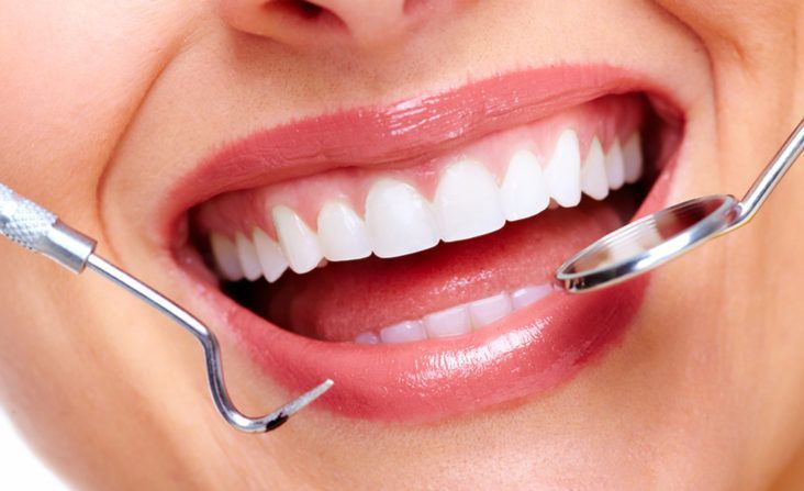 A close-up of a person's smiling mouth during a dental checkup, with a dental mirror and explorer tool visible.