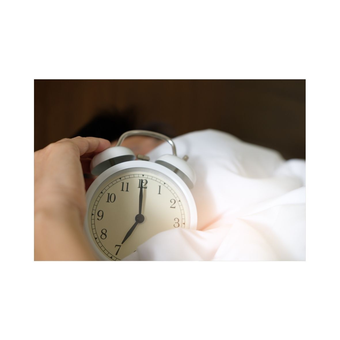A person's hand reaching for a white alarm clock set at 7:00 am, emerging from white bedding.