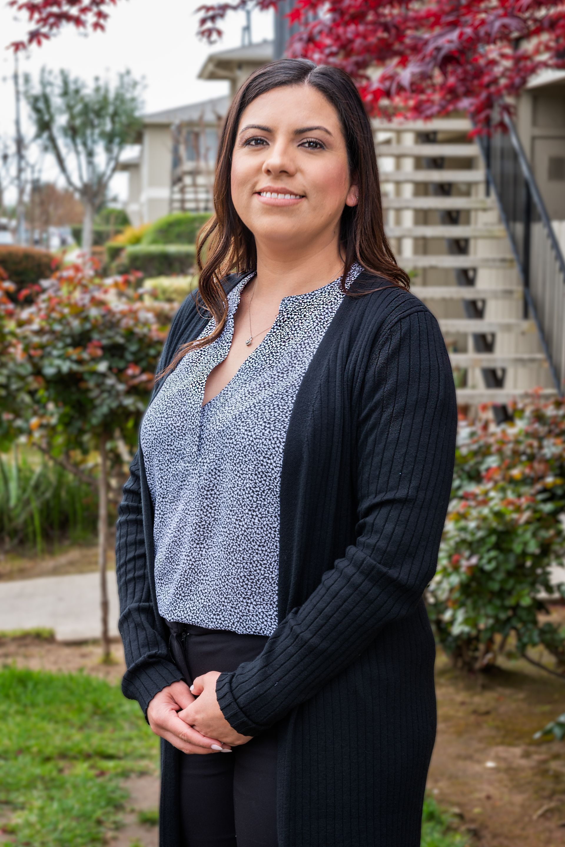 Woman in black cardigan and patterned blouse standing outside, smiling.