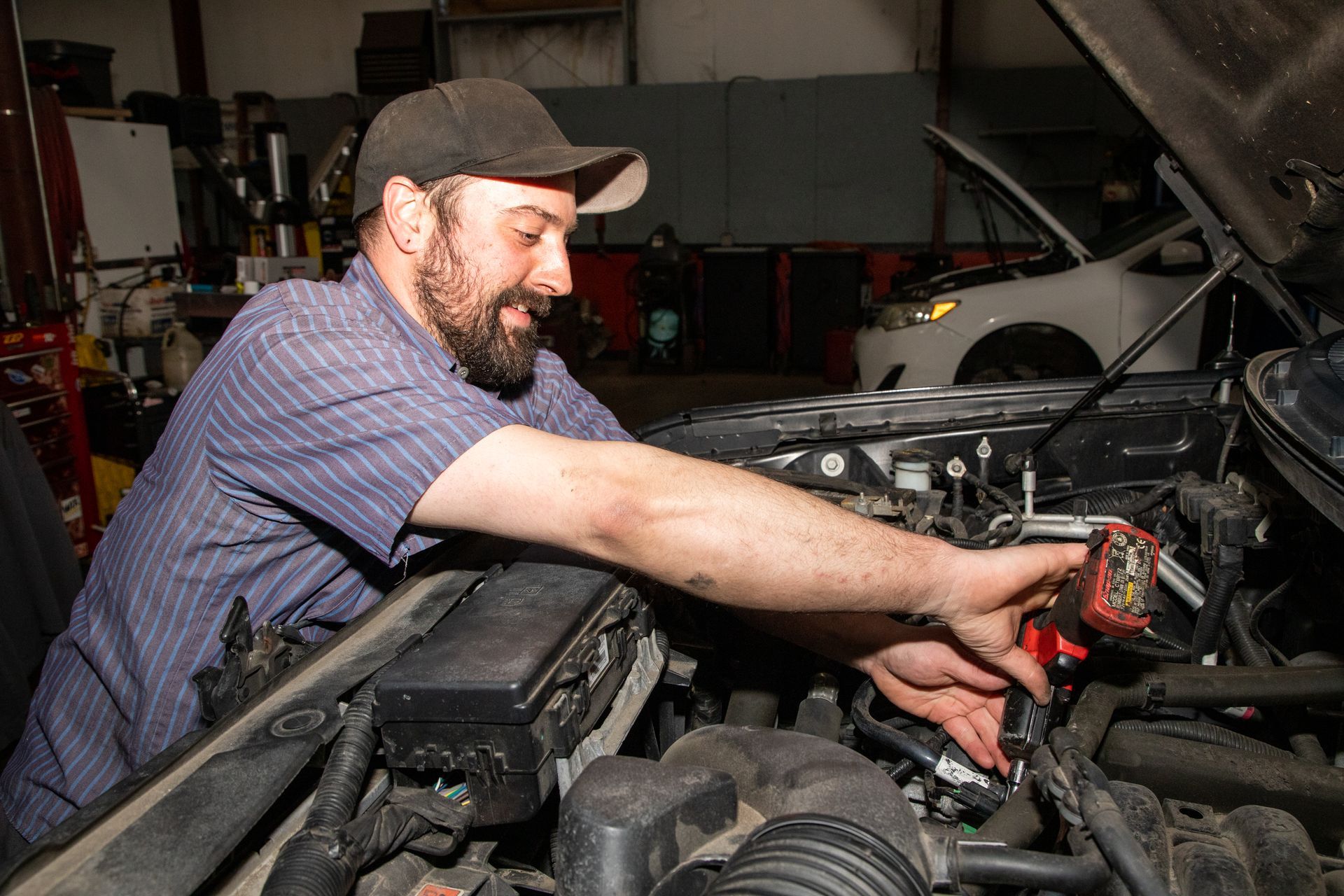 Mechanic working under an open car hood in a garage, using red pliers on engine parts