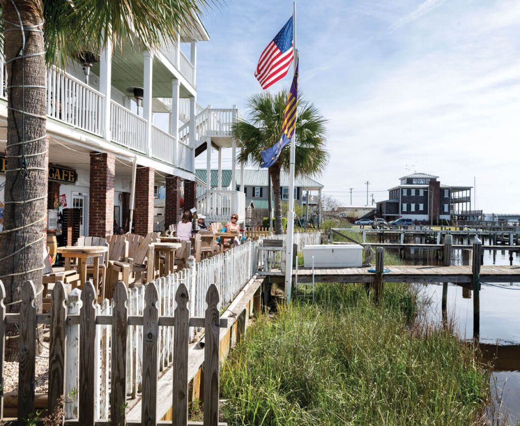 Restaurant with outdoor seating, waterfront view. American flag waves. White fence, water, building with balconies.