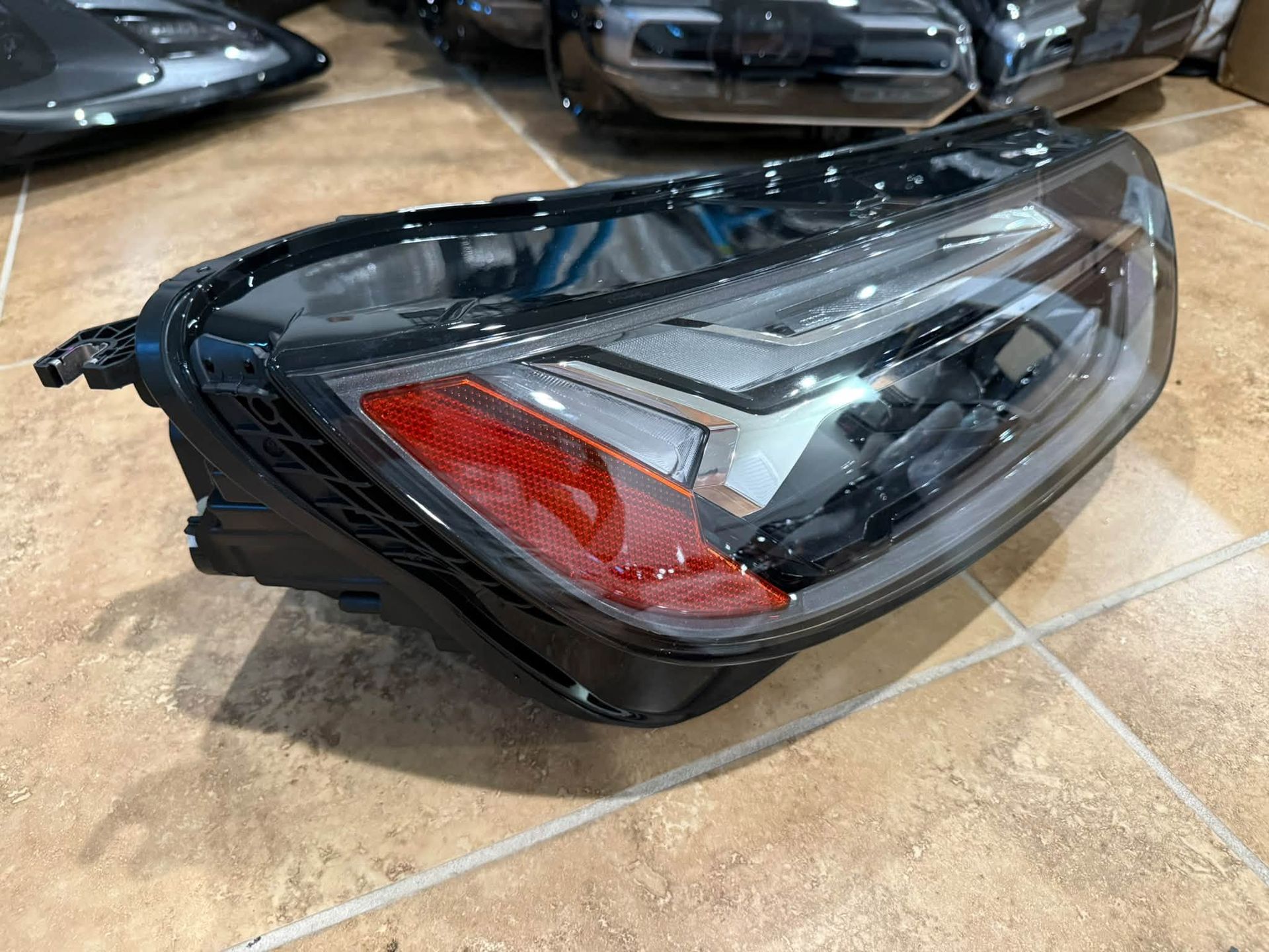 Black car headlight assembly with red accent, resting on a tiled floor near other car parts