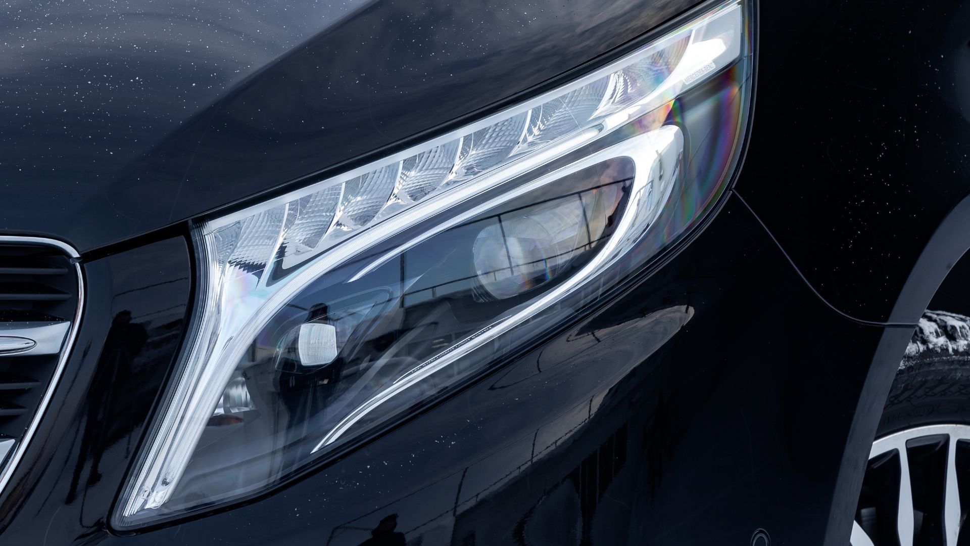 Close-up of a black car’s front headlight and grille, showing sleek chrome detailing.