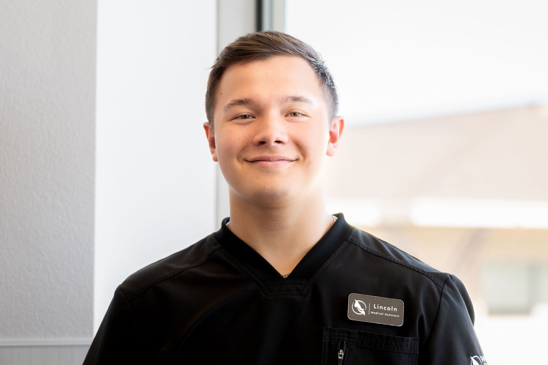 Dennis the Medical Assistant is in a black scrub top is smiling with his arms crossed.