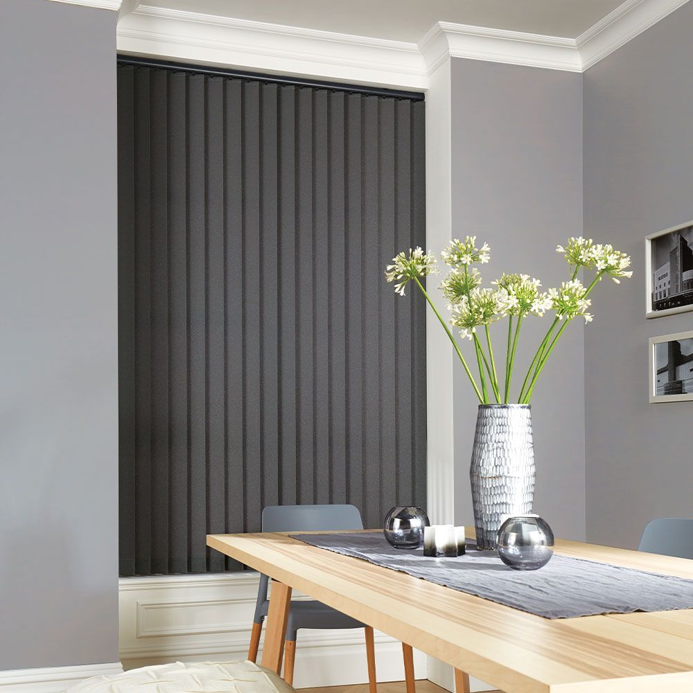 Black vertical blinds in a gray dining room, wooden table, silver vase with white flowers, gray walls.