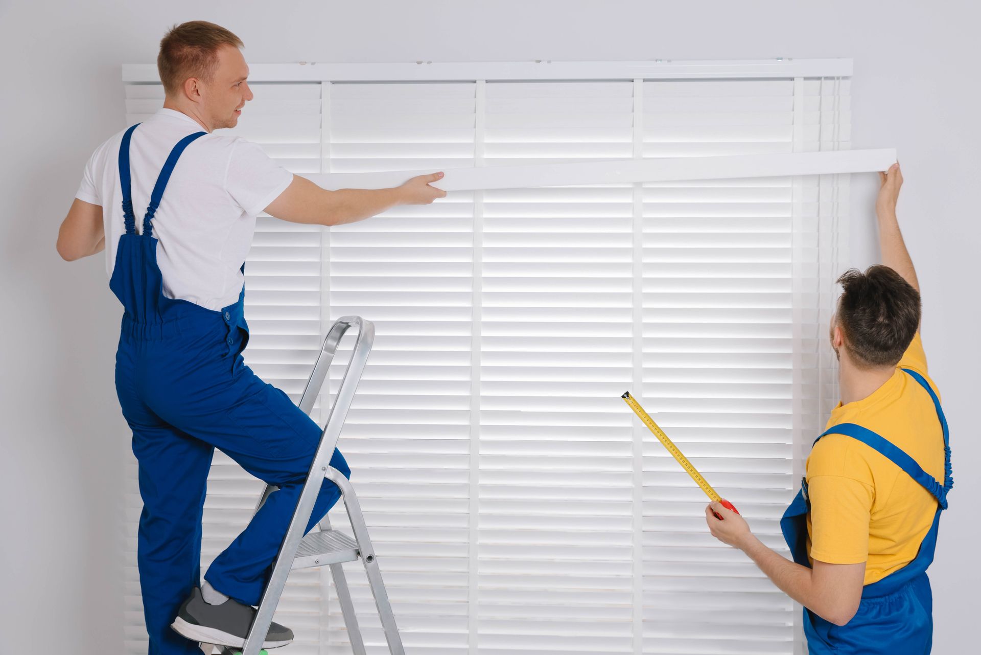 Two workers installing white blinds on a window. One on a ladder, measuring; the other holding the blind.