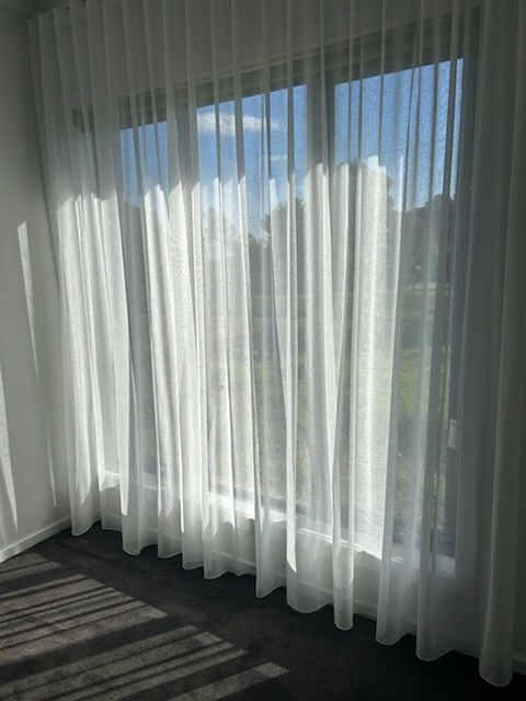 Sheer white curtains hanging in front of a window; sunlight streams through, casting shadows.