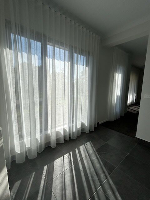 Room with sheer white curtains, sunlight streaming through a large window onto gray tiled floor.