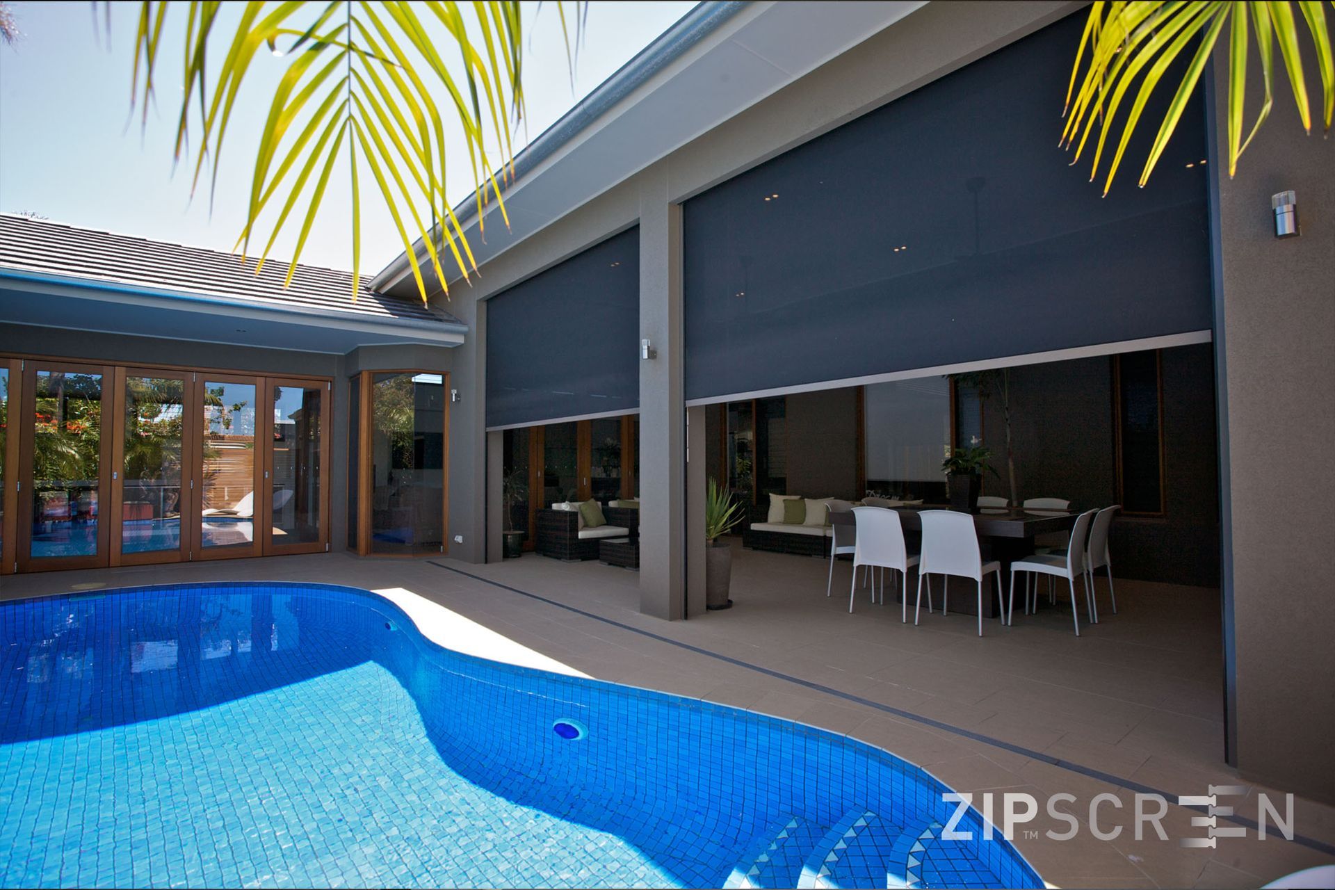 Poolside patio with dark gray retractable screens, open onto a dining area. Blue pool and palm leaves visible.