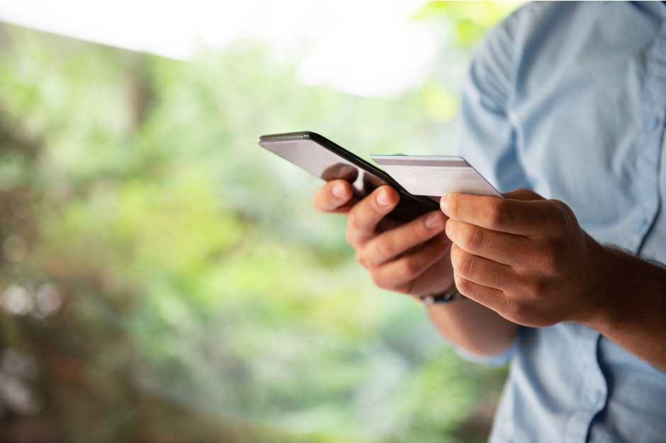 Person holding a credit card and smartphone near a window, likely making an online purchase.