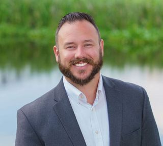Man in a grey blazer smiling, standing in front of a body of water.