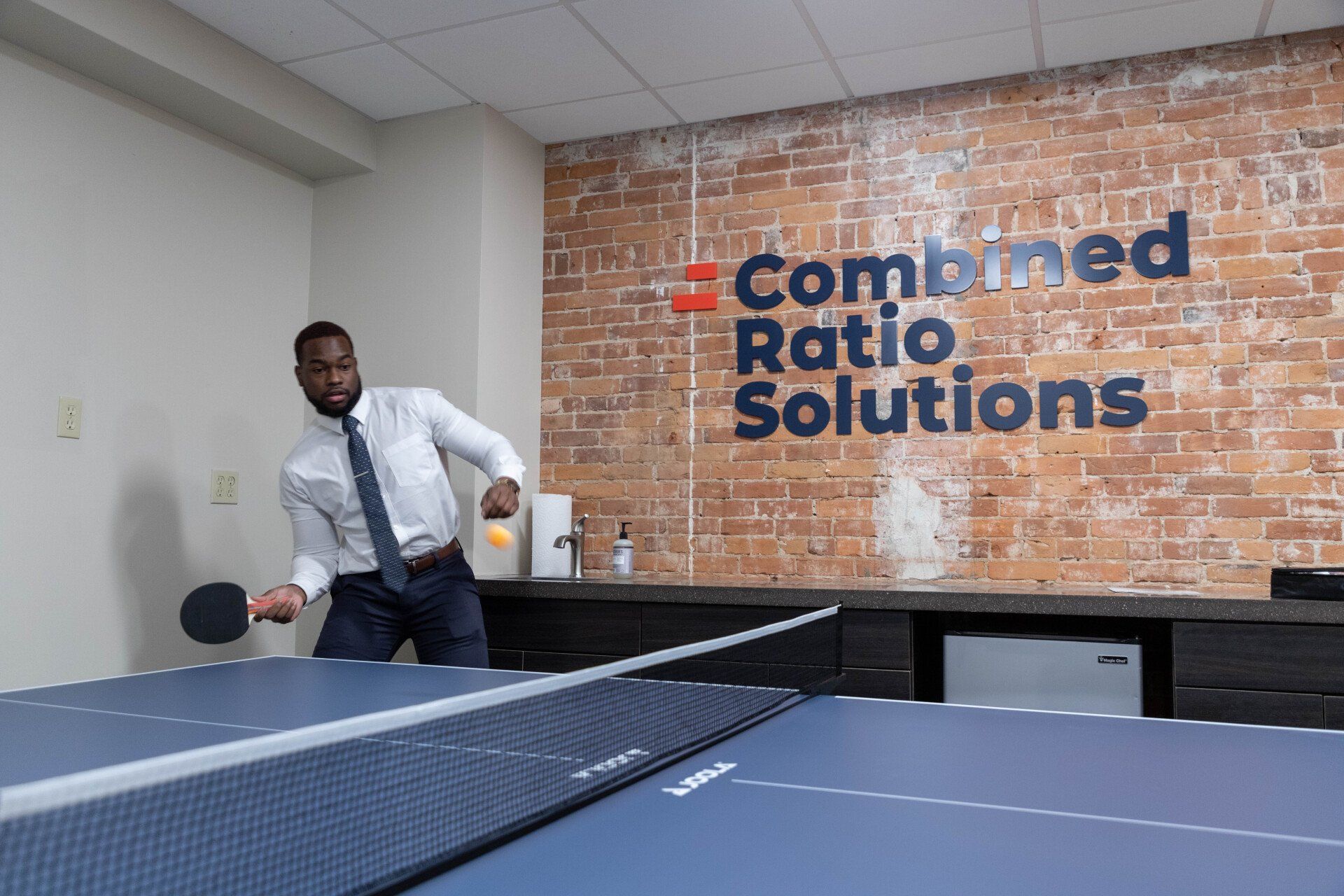 A man is playing ping pong in front of a brick wall that says combined ratio solutions.