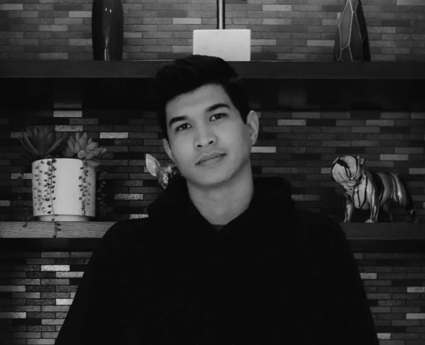 A black and white photo of a young man standing in front of a brick wall.