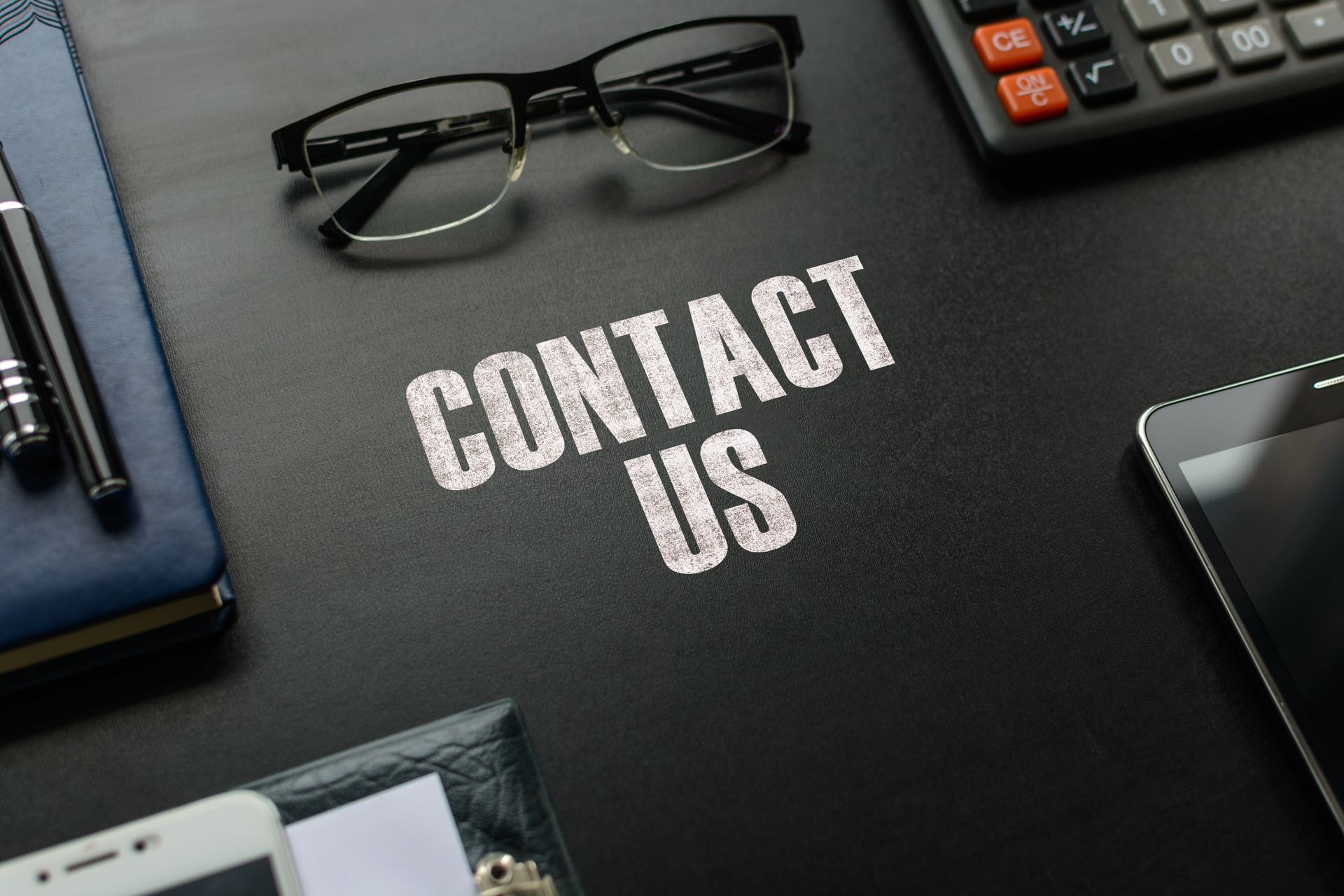 Contact Us text on black desk surrounded by glasses, phone, notebook, calculator, and pen.